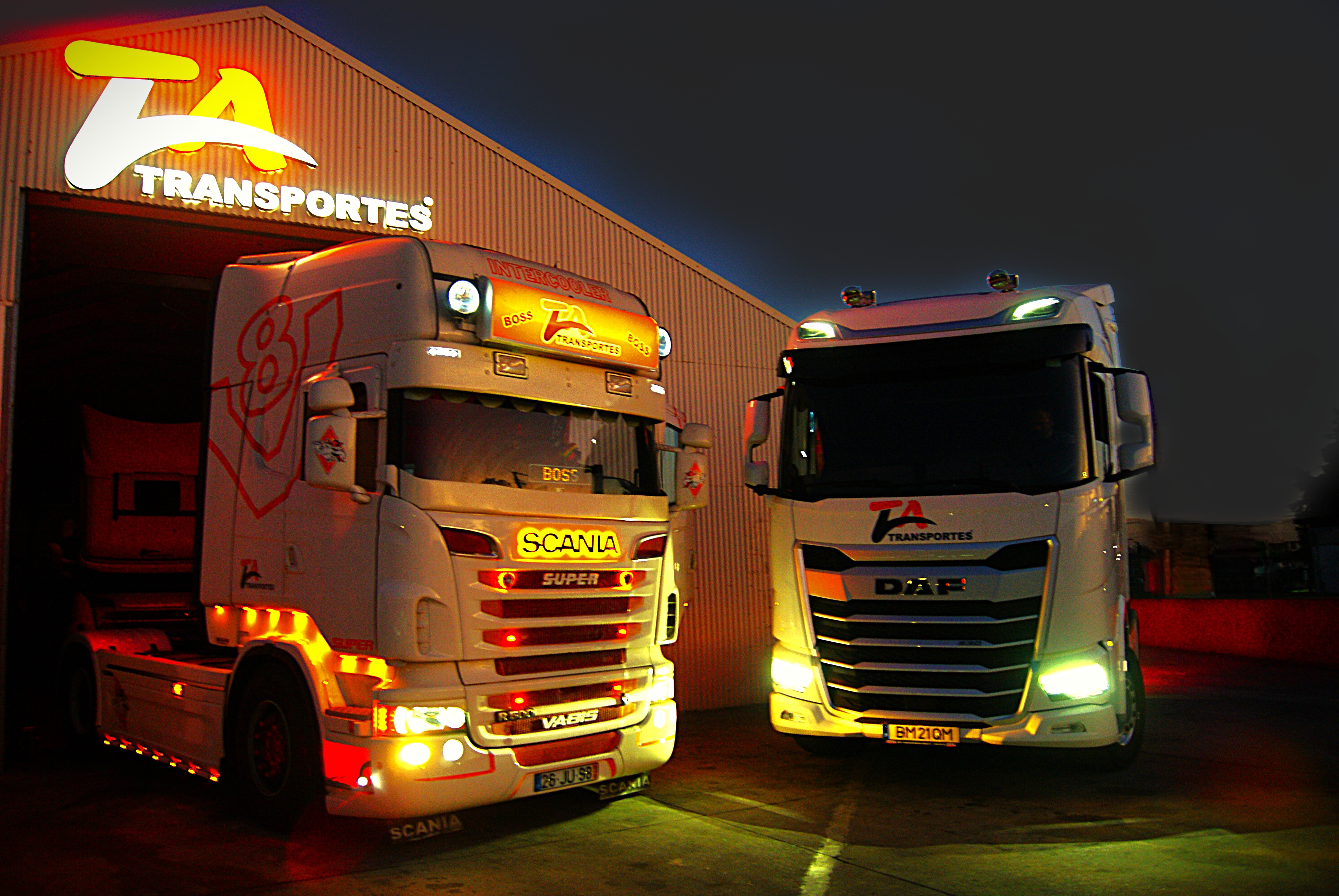 Imagem de dois camiões de noite à frente do armazém da TaTransportes que tem o logotipo LED em cima ligado.