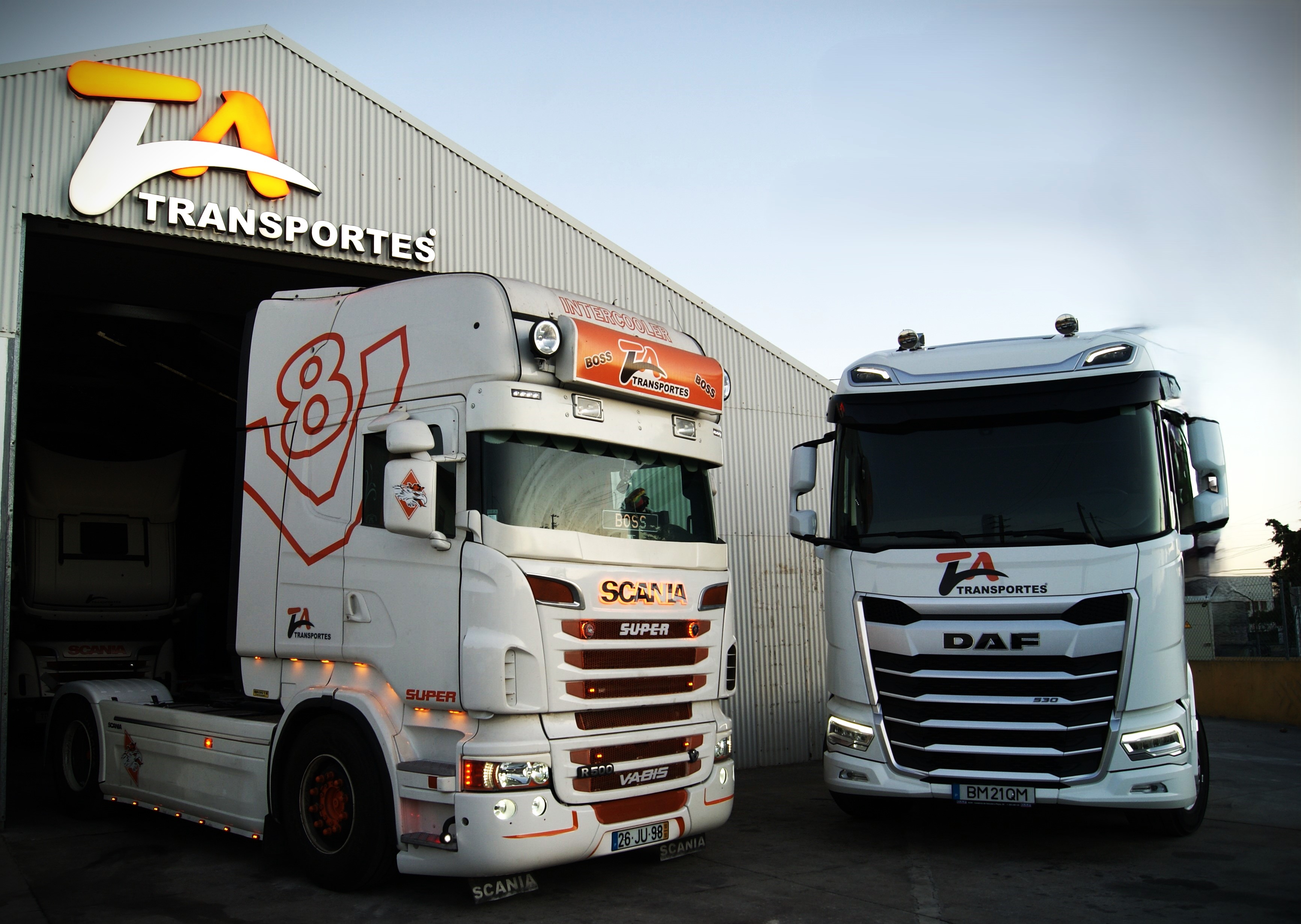 Imagem de dois camiões de noite à frente do armazém da TaTransportes que tem o logotipo LED em cima ligado.